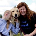 Young girl with hearing aid hugs yellow lab service dog as the girls mother smiles on.