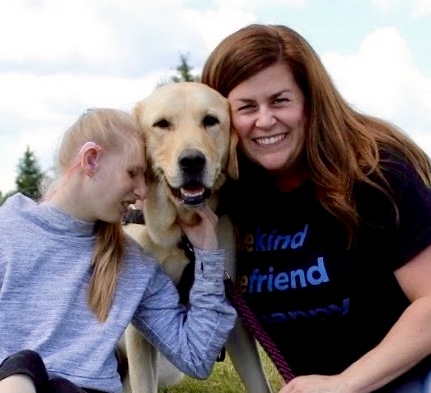 Young girl with hearing aid hugs yellow lab service dog as the girls mother smiles on.