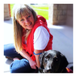 A blonde woman smiles, sitting with a service dog in training.