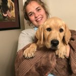 A woman smiles at the camera while holding a golden retriever puppy wrapped in a brown towel. A framed painting of a dog is on the wall behind them.