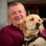 Man in maroon shirt holds a yellow Labrador PawPADs service dog on his lap.