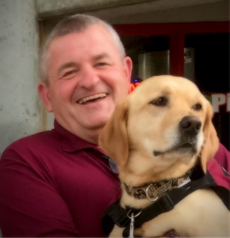 Man in maroon shirt holds a yellow Labrador PawPADs service dog on his lap.