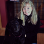 A blonde woman smiles, sitting with a service dog in training.